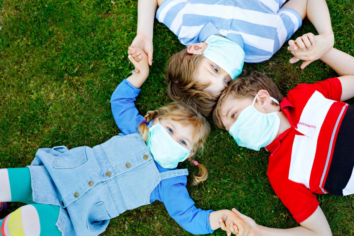 Three masked children lying on the grass holding hands